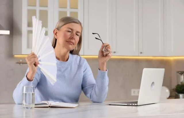 woman waving hand fan to cool herself during hot flash for article online menopause care